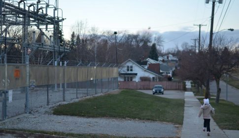 A young child running down a street in Flint, MI, past an electric transformer station