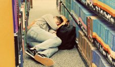 A student sleeping in the aisle of a library with his knapsack