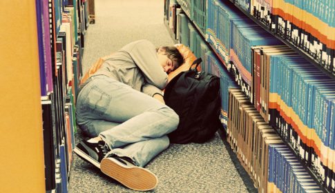 A student sleeping in the aisle of a library with his knapsack