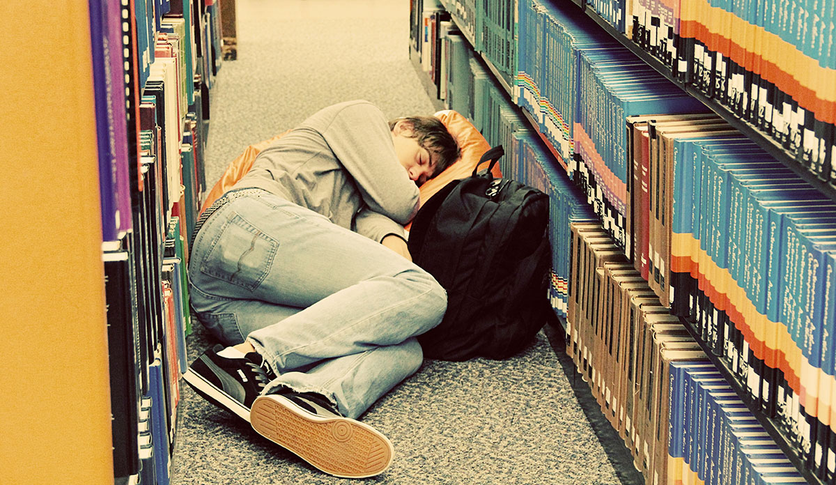 A student sleeping in the aisle of a library with his knapsack