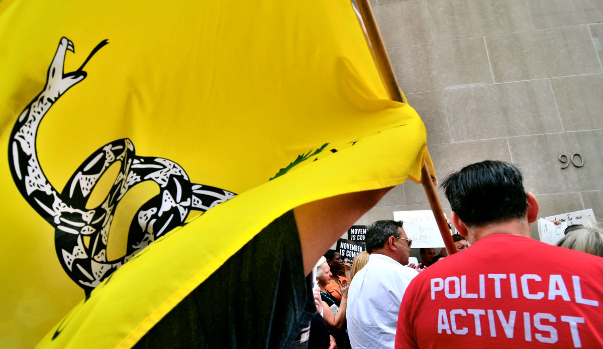 A man in a red shirt with "Political Activist" on the back, holding a yellow Don't Tread on Me Tea Party flag