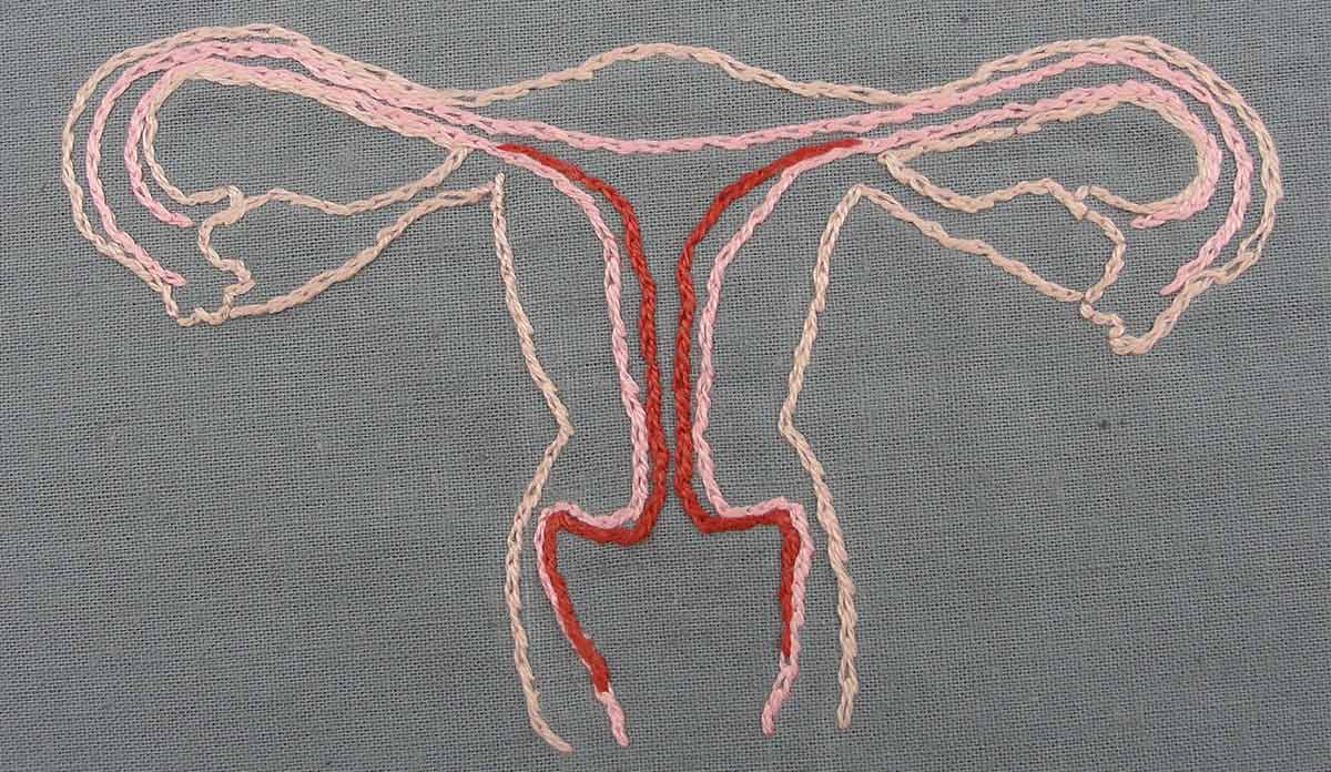 A needlepoint of a uterus in pink and red thread on a grey fabric background