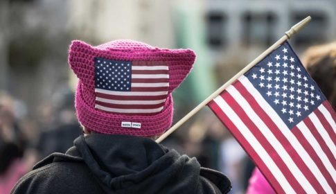 The back of the head of a person carrying an American flag, wearing a pink knitted "pussy hat" with an American flag on it