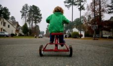 A child in a green jacket on a red tricycle pedals down the middle of a town street