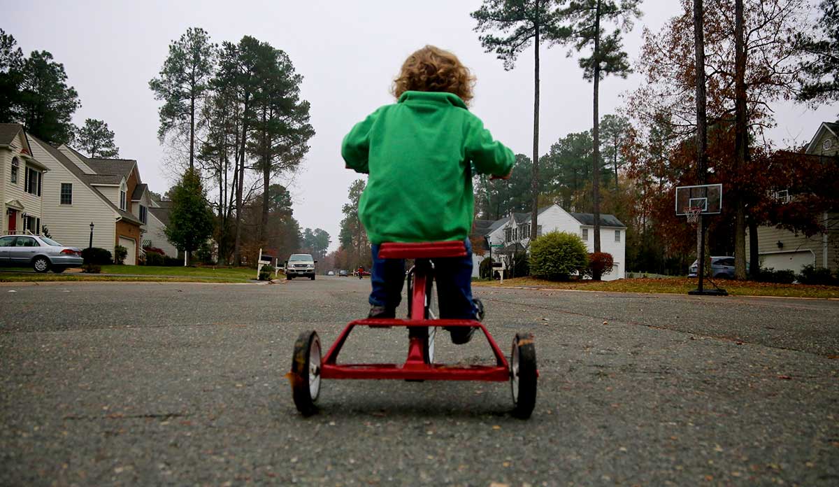 A child in a green jacket on a red tricycle pedals down the middle of a town street