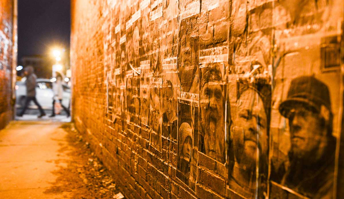 Night shot of alley with photos of people's faces wheatpasted on the bricks