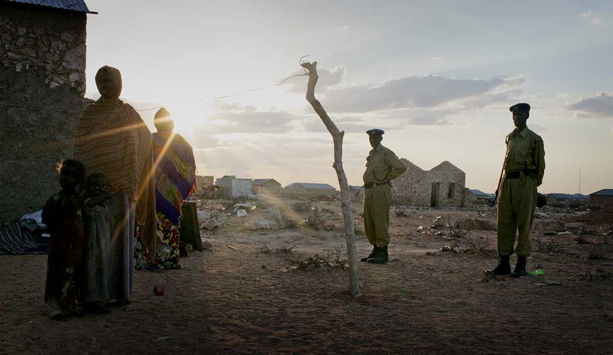 A woman and her young children stand in the evening light at an IDP settlement in South Galkayo, Somalia with armed police standing by.