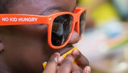 Child eating and orange, wearing orange sunglasses that say No Kid Hungry