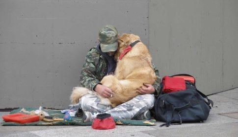 Man sitting on sidewalk with his belongings, with a dog in his lap