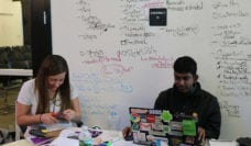 Two hackathon participants sitting at a table in front of a whiteboard with ideas