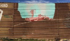 Mural painted on border wall of a gap with a person lying on their back, reaching toward blue sky