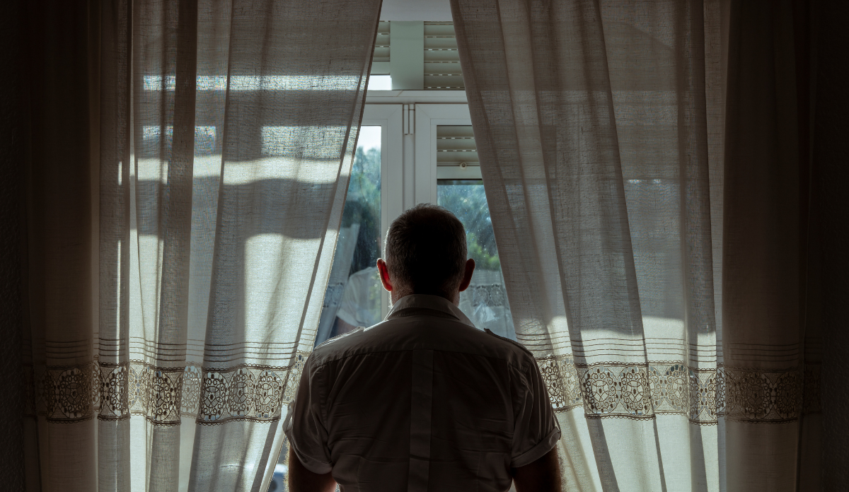 Rear view of adult man standing beside window with sunlight and shadow
