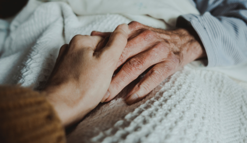 Close-up of a young woman's hand holding the hand of an older man lying on a bed. Caregiving concept